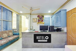 Un mostrador de recepción con un cartel que dice 'HOTELO Company-Serviced', un ventilador de techo, un reloj, un monitor de computadora e impresora.