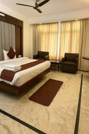 A hotel room with a large bed, two armchairs, a ceiling fan, and a view of the outside through curtains. - The Grand Valley (Mussoorie)