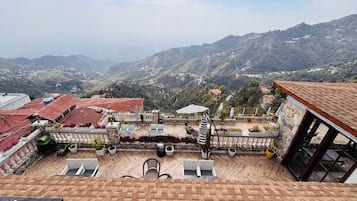 Una terraza con mesas al aire libre, vistas a un paisaje montañoso y un edificio con tejado de tejas rojas.