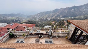 A terrace with outdoor seating, a view of a mountainous landscape, and a building with a red-tiled roof. - The Grand Valley (Mussoorie)