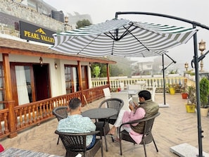 Zona d'estar exterior amb cadires de vimet, un parasol rayat, una barana de fusta i un cartell que diu 'PEARL VALLEY'.