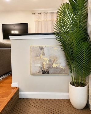 A living room with a flat-screen TV, a framed abstract painting, a potted plant, and a patterned rug.