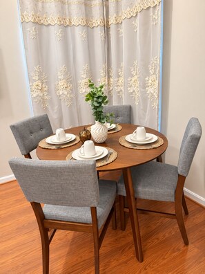 A round dining table with four grey upholstered chairs, a white vase with a green plant, and white plates and cups set on woven placemats.