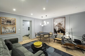 Ein modernes Wohnzimmer mit einer Couch, einem Esstisch mit Stühlen, einem Couchtisch mit Obstschale und einem großen Gemälde an der Wand.