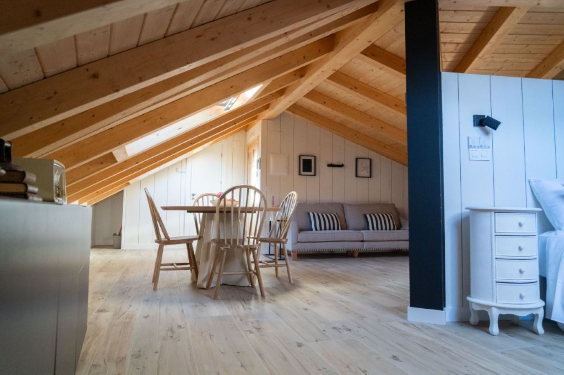 An attic room with a wooden ceiling, a dining table with chairs, a sofa, and a white dresser.