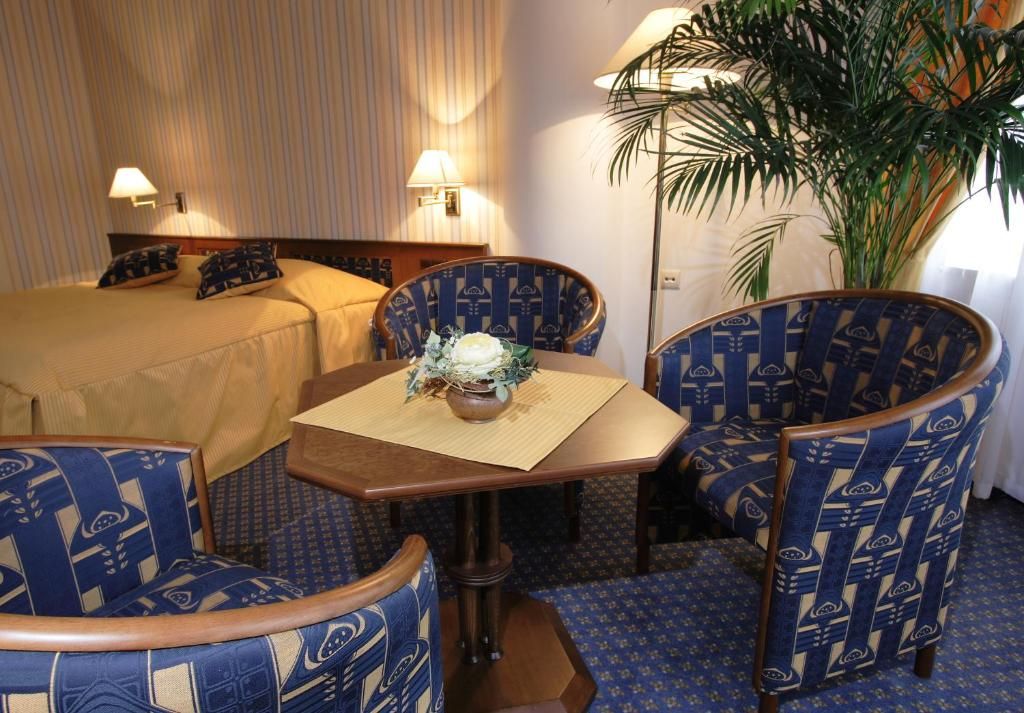 A hotel room with a bed, two chairs, a small table with a vase, and a large plant.
