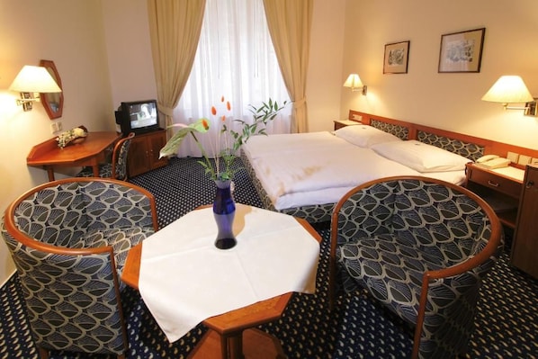Una habitación de hotel con cama, dos sillones, una mesita, televisor y un jarrón con flores.