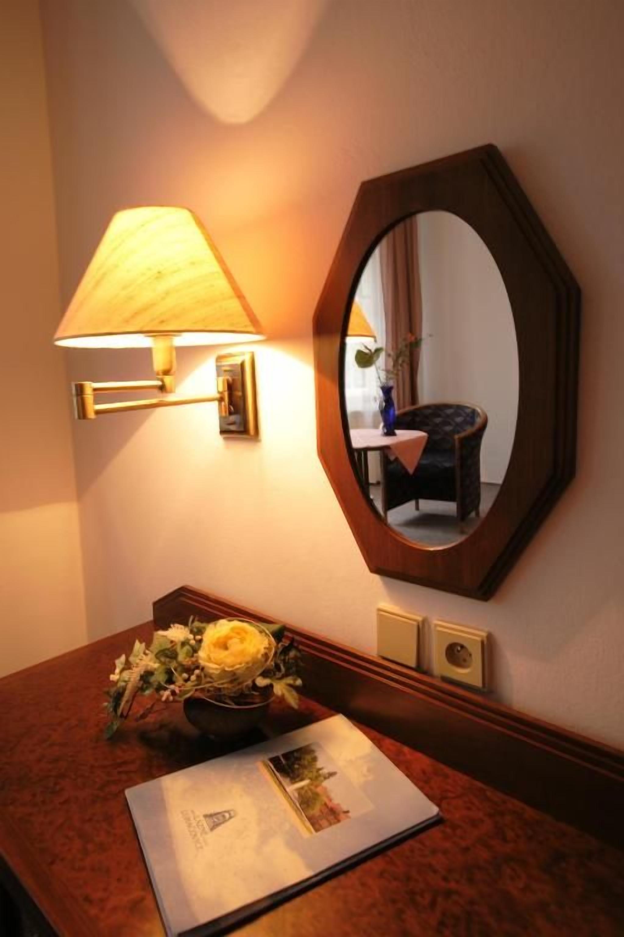 A hotel room with a wooden desk, a lamp, a mirror, and a vase with flowers.