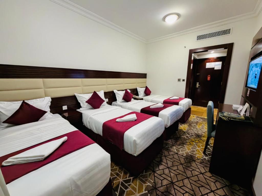 A hotel room with four beds, each with white linens and red accents, a patterned carpet, and a television on the right.