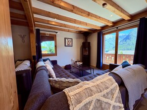A cozy living room with wooden beams, a fireplace, and a large window offering a mountain view. - Chalet Doux Abri, sleeps 12, hot tub, sauna (Morzine)