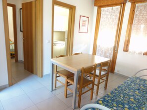 A room with a wooden dining table and chairs, a bed with a patterned bedspread, a window with lace curtains, and a framed certificate on the wall. - Apartment With Terrace in Village With Pools (San Michele al Tagliamento)