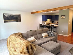 A living room with a sectional sofa, a pet dog, and a view into the kitchen area. - SlopeTown Retreat - Big Sky Meadow (Big Sky)