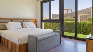 Quarto com cama grande, cabeceira de madeira e vista para uma parede de pedra e vegetação lá fora.