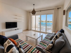 A modern living room with a flat-screen TV, a sofa set, a coffee table, and a view of a pool and buildings through large glass doors. - Vesta - Luxury APT - 2BR - Joubal (El Gouna)