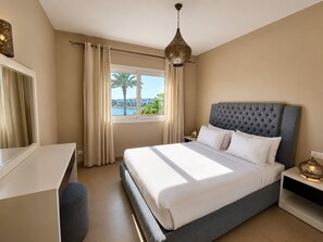 A bedroom with a bed, a desk, a chair, a nightstand, a lamp, and a view of the outdoors. - Vesta - Luxury APT - 2BR - Joubal (El Gouna)