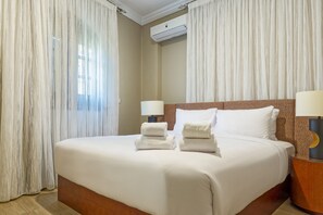 A hotel room with a large bed, two bedside lamps, a headboard, and a wall-mounted air conditioner. - Luxury APT - 2BR - South Teseen I (New Cairo)