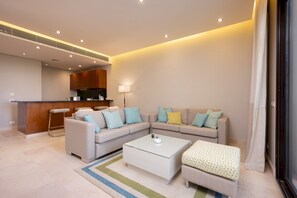 Ein modernes Wohnzimmer mit einer Couch, einem Couchtisch und einem kleinen Hocker. Im Hintergrund befindet sich ein Küchenbereich mit einer Bar und Hockern.