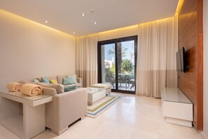 Ein modernes Wohnzimmer mit einer Couch, einem Couchtisch und einem Fernseher. Ein Glastisch mit Handtüchern und ein großes Fenster mit Vorhängen.