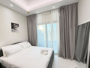 A modern hotel room with a large bed, a window with curtains, and a ceiling light. - Vesta - Luxury Studio - Hera Tower (Dubai)