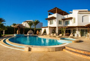 Ein Poolbereich mit einem runden Pool, Liegestühlen und einem Gebäude mit heller Fassade und Holzpergolen.