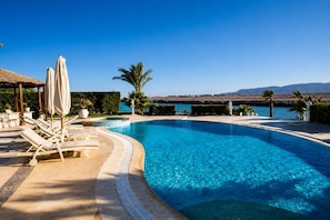 Ein klares, blaues Schwimmbecken mit Liegestühlen und Sonnenschirmen, umgeben von Grünpflanzen und Blick auf das Meer.