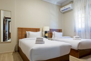 A hotel room with two beds, a nightstand with a lamp, an air conditioning unit, and a window with curtains. - Luxury APT - 3BR - South Teseen I (New Cairo)