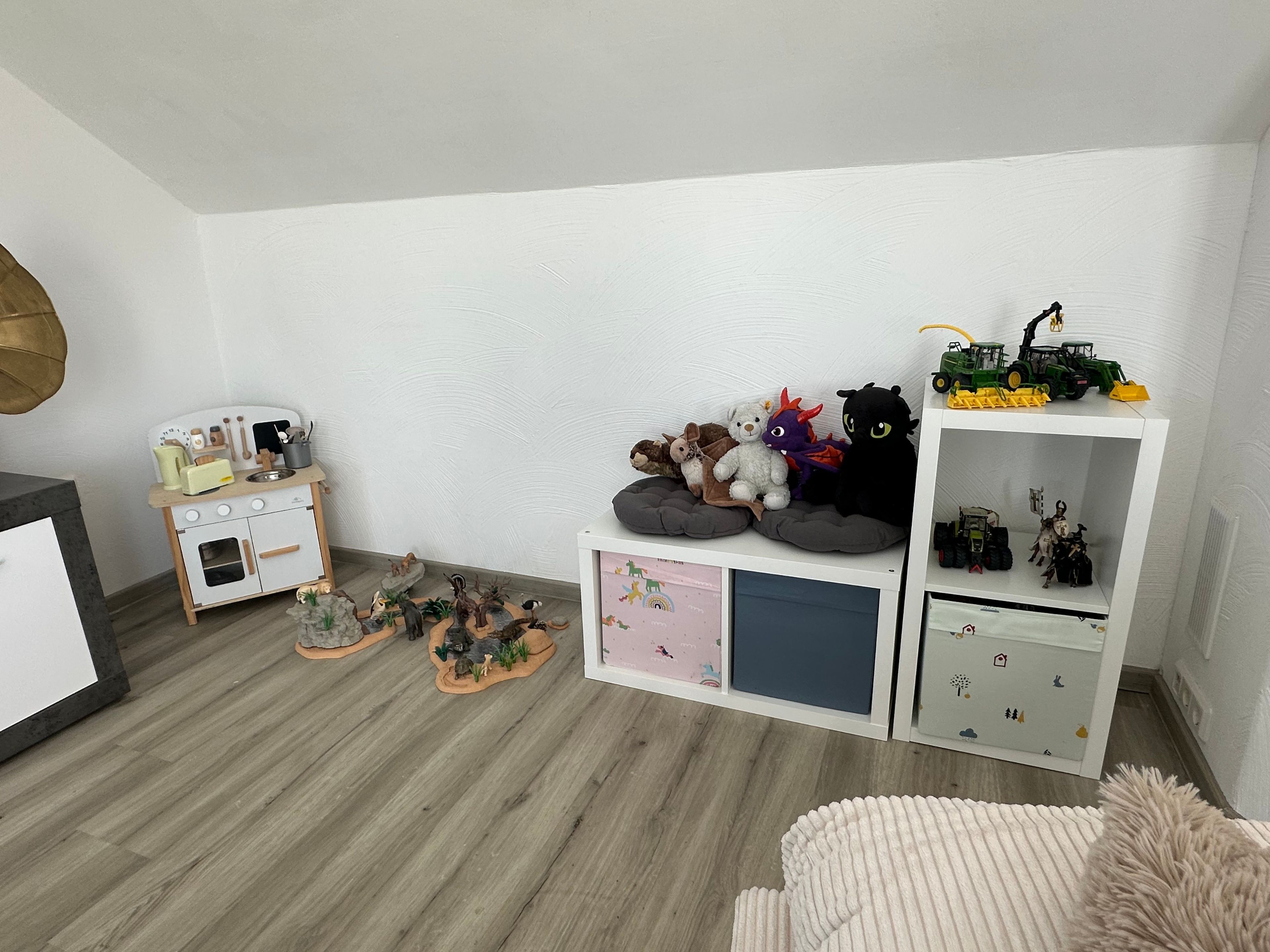 Ein Kinderzimmer mit weißer Wand, einer Spielküche und einem Regal voller Spielzeug und Figuren.