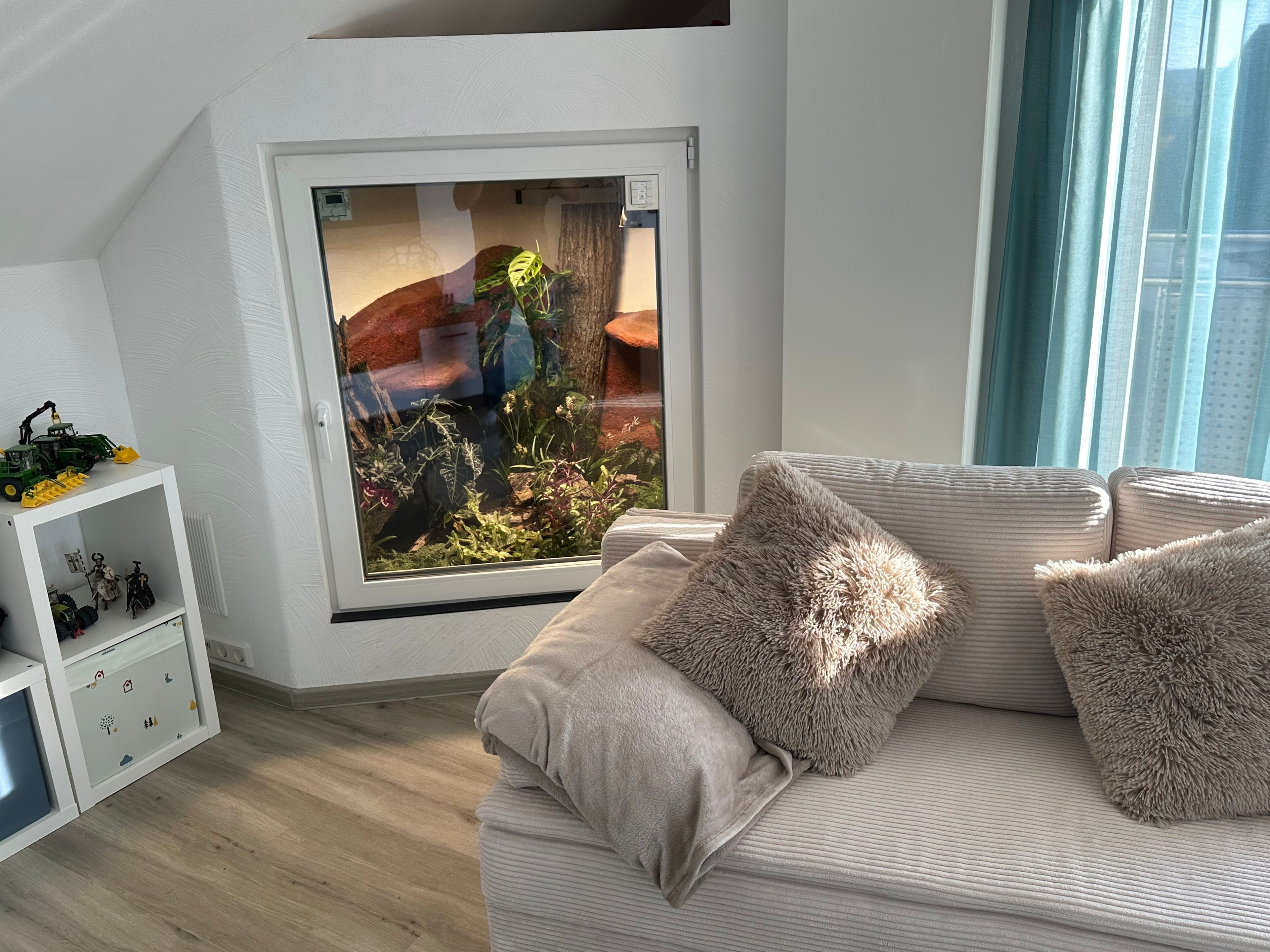 Una habitación con un gran ventanal, una pared blanca, un sofá con cojines mullidos y una pequeña estantería con figuritas.