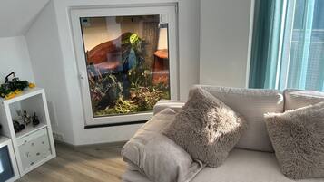 Una habitación con un gran ventanal, una pared blanca, un sofá con cojines mullidos y una pequeña estantería con figuritas.