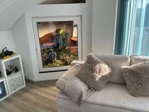 A room with a large window, a white wall, a sofa with fluffy cushions, and a small shelf with figurines.
