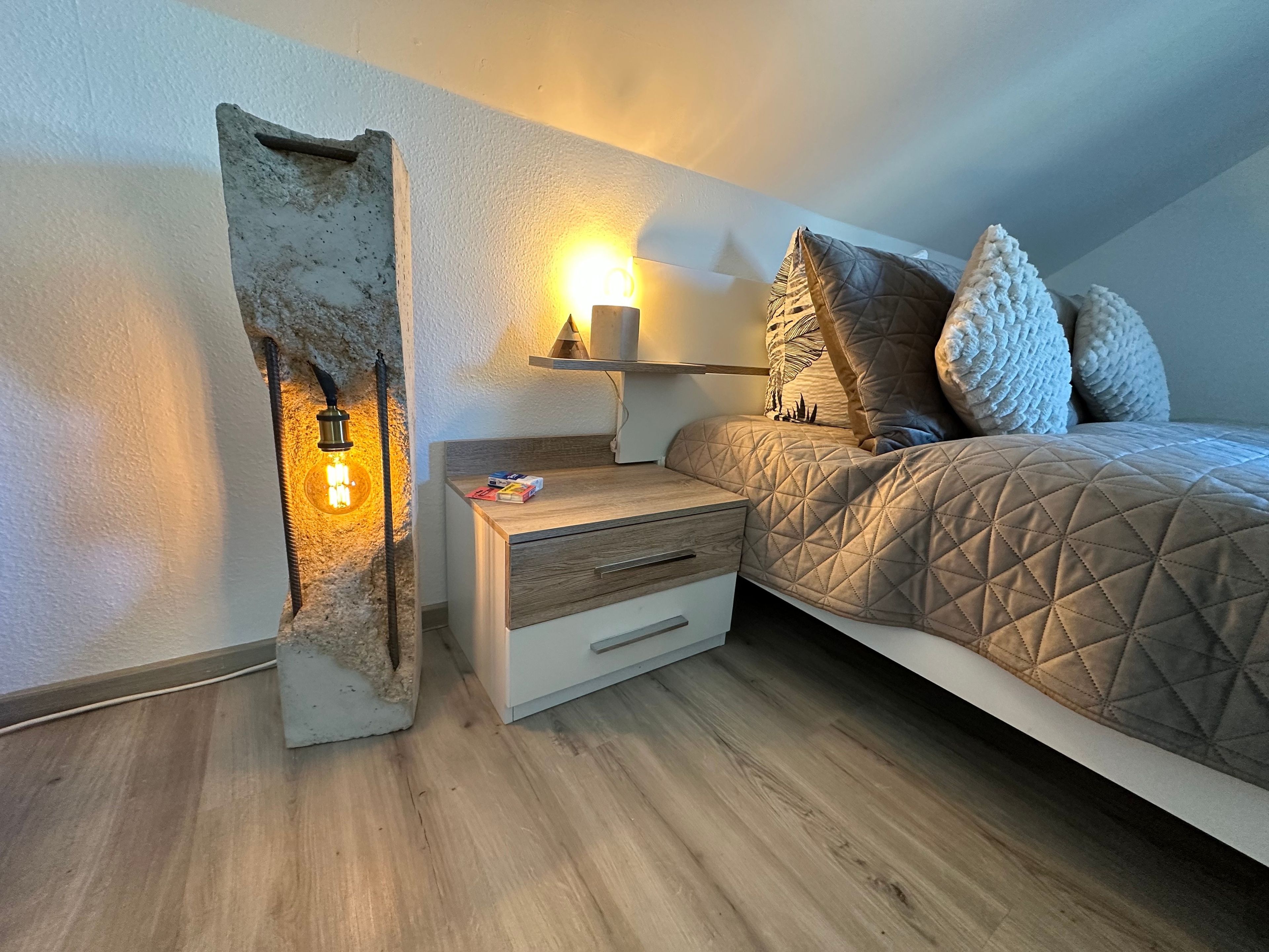 Quarto com cama, mesa de cabeceira, luminária e prateleira na parede.
