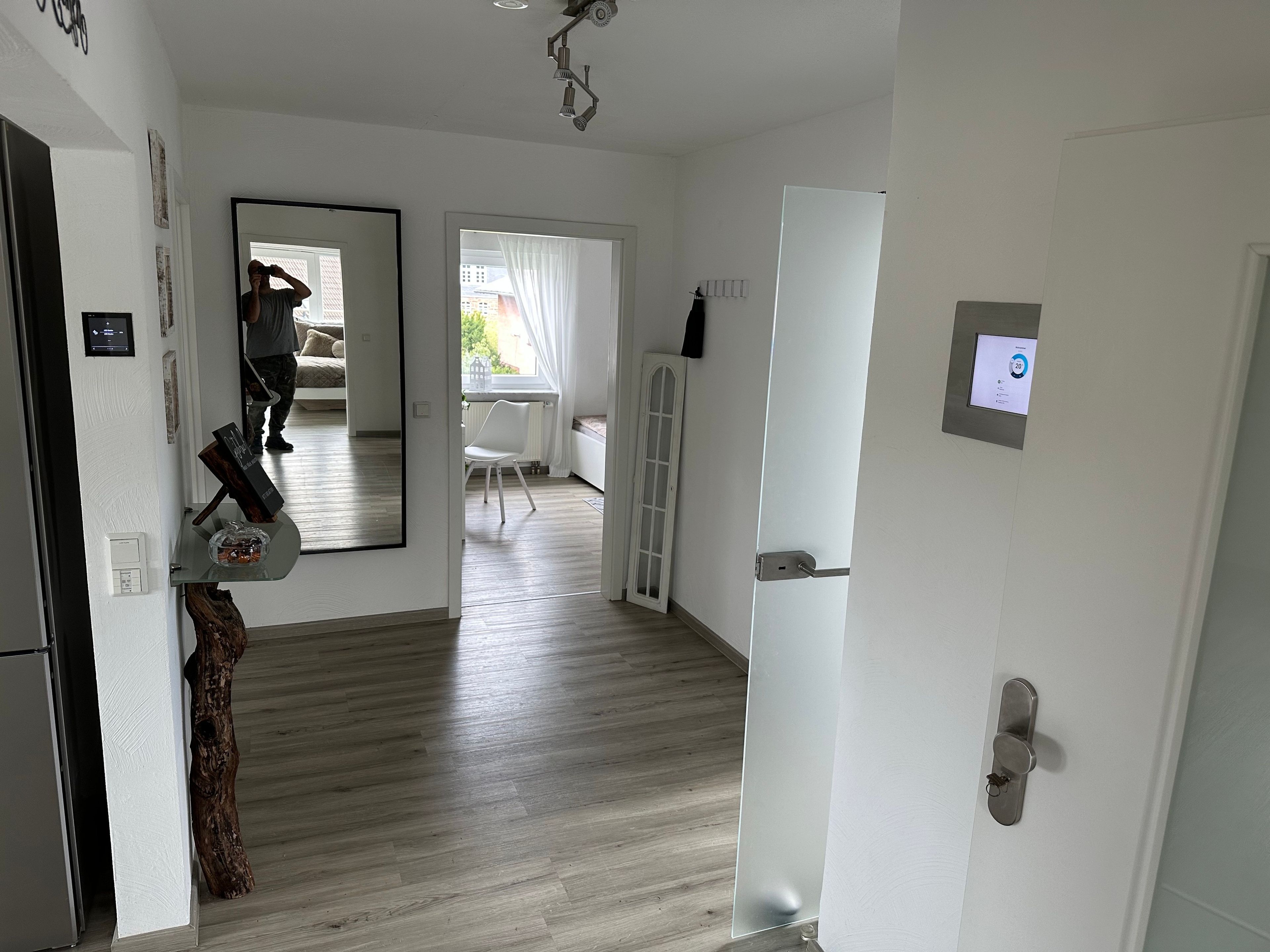 Ein modernes Interieur mit Holzboden, einer weißen Tür mit digitaler Anzeige und einem Spiegel, der einen Raum mit Fenster und Stuhl reflektiert.