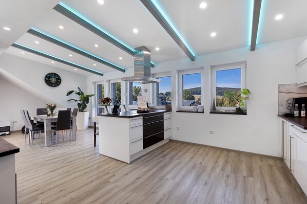 A modern kitchen with a central island, wooden flooring, and large windows offering a view of the outdoors.