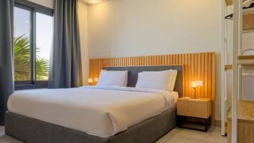 Habitación de hotel con una cama grande, cabecera de madera, mesita de noche con lámpara y vistas a palmeras a través de la ventana.