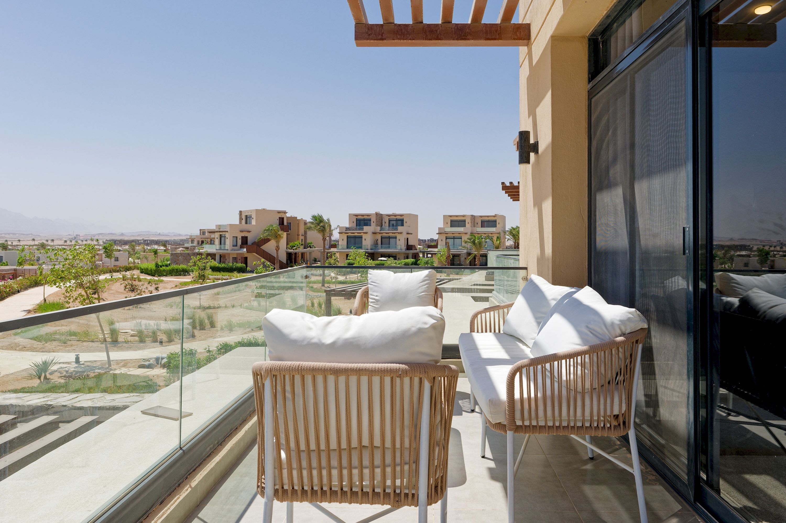 Un balcón con mobiliario de mimbre y barandilla de vidrio con vistas a un área residencial.