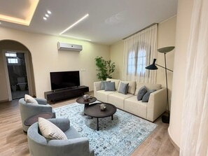 Ein modernes Wohnzimmer mit Flachbildfernseher, einem beigefarbenen Sofa, zwei Sesseln, einem Couchtisch und einer Stehlampe.