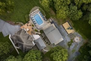 An aerial view of a property with a swimming pool, a covered patio, and a large building with a unique roof structure. - Spa House, Sauna, Pool, Arcade, Theater, Pickleball Court, & More! (Mount Union)