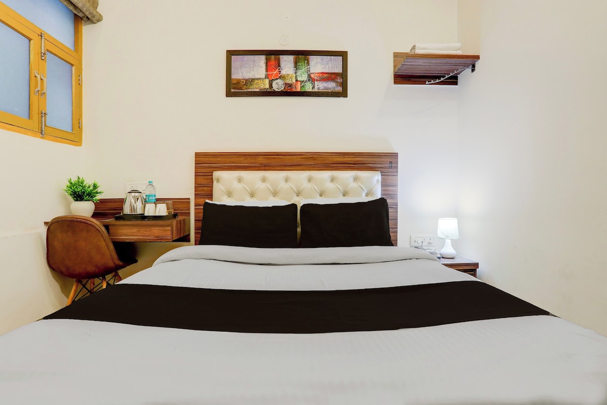 Habitación de hotel con cama, mesita de noche, lámpara, estante en la pared, ventana con marco de madera y una pequeña planta en maceta.