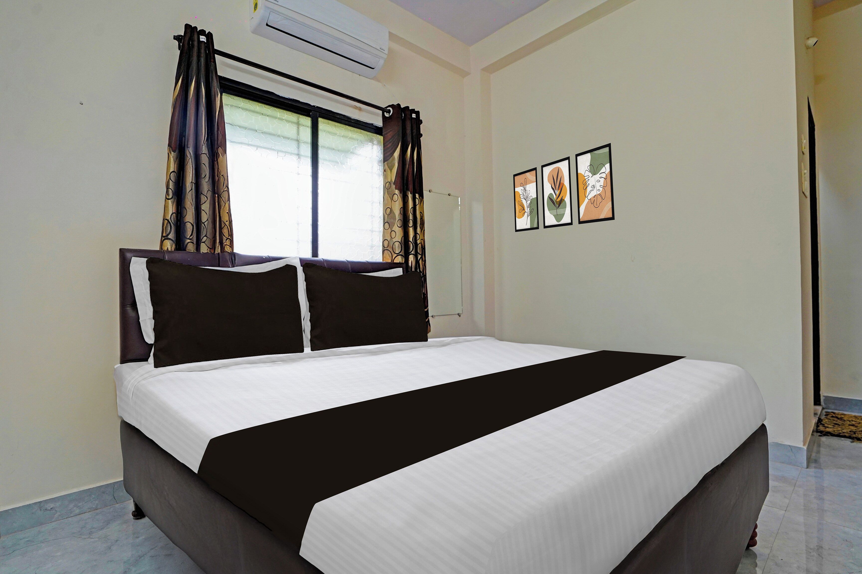 A hotel room with a bed, curtains, an air conditioning unit, and two framed artworks on the wall.
