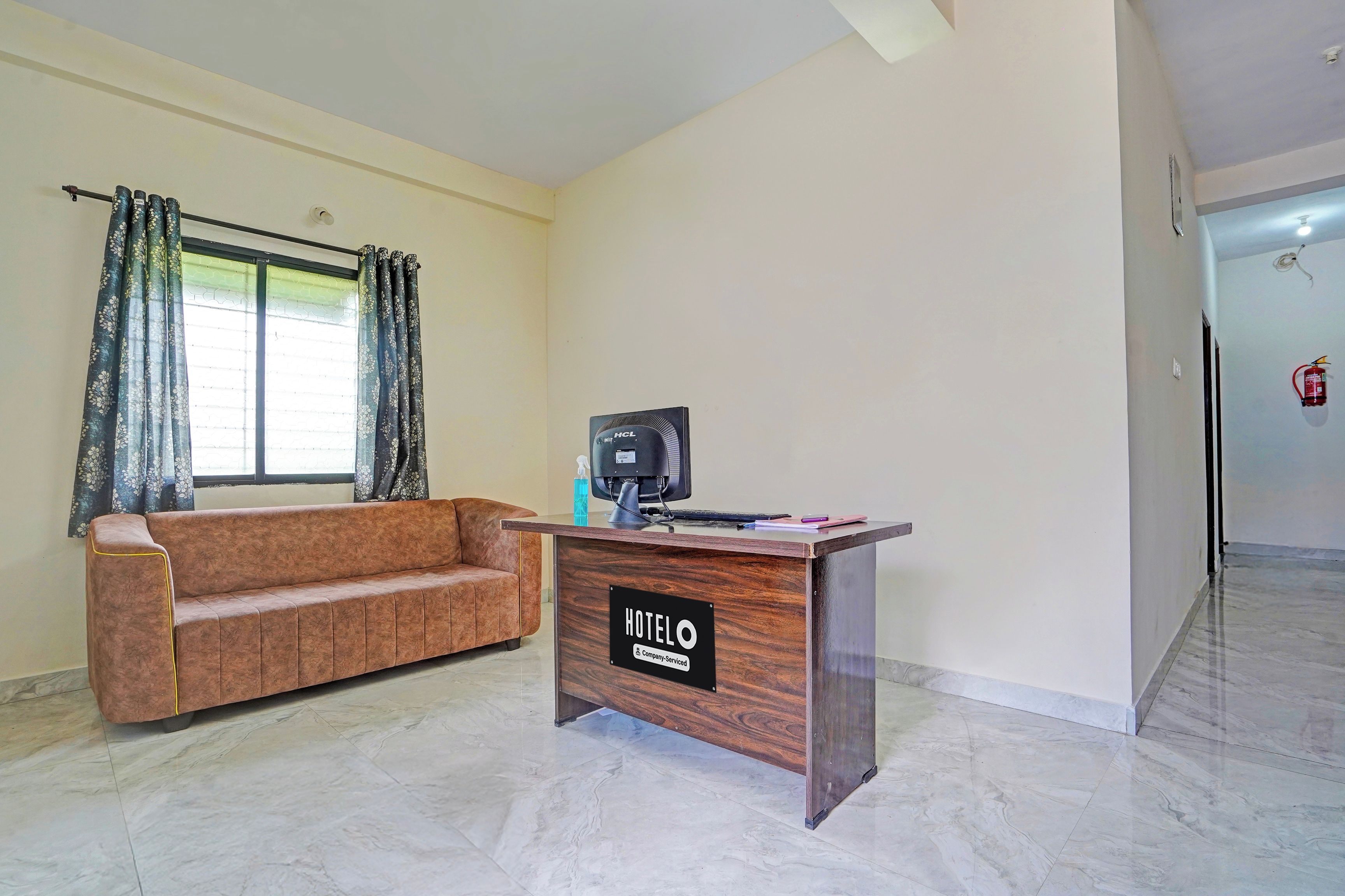 A hotel reception area with a wooden desk, a sofa, a window with curtains, and a fire extinguisher on the wall.