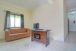 A hotel reception area with a wooden desk, a sofa, a window with curtains, and a fire extinguisher on the wall.