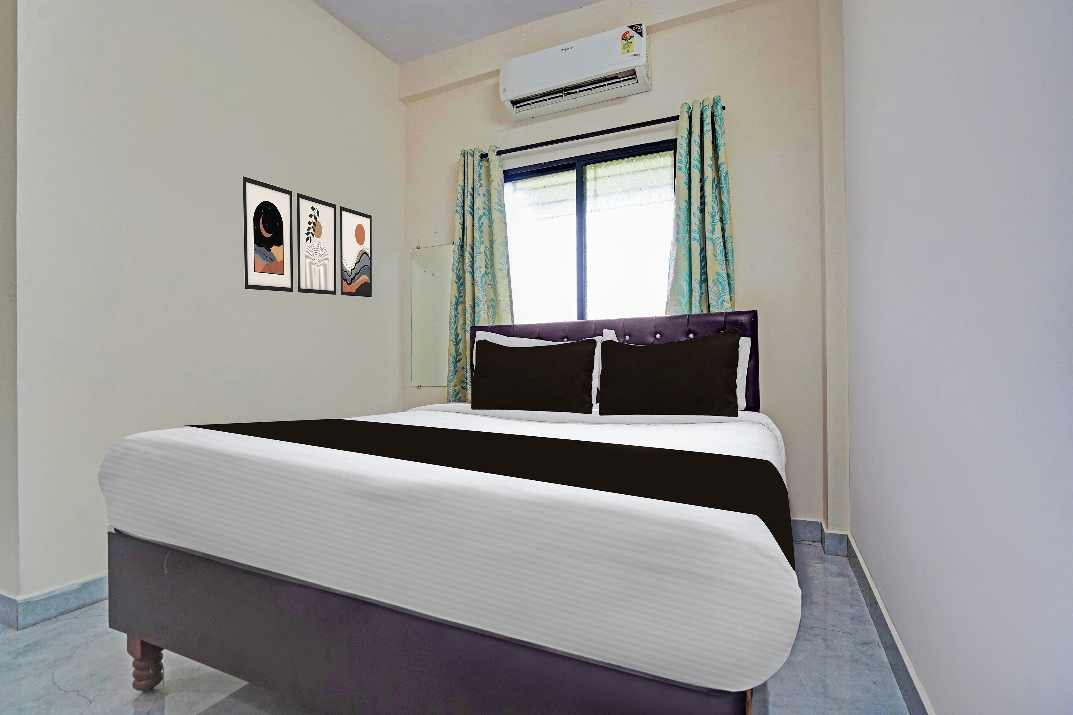Una habitación de hotel con cama, aire acondicionado, ventana con cortinas y cuadros en la pared.