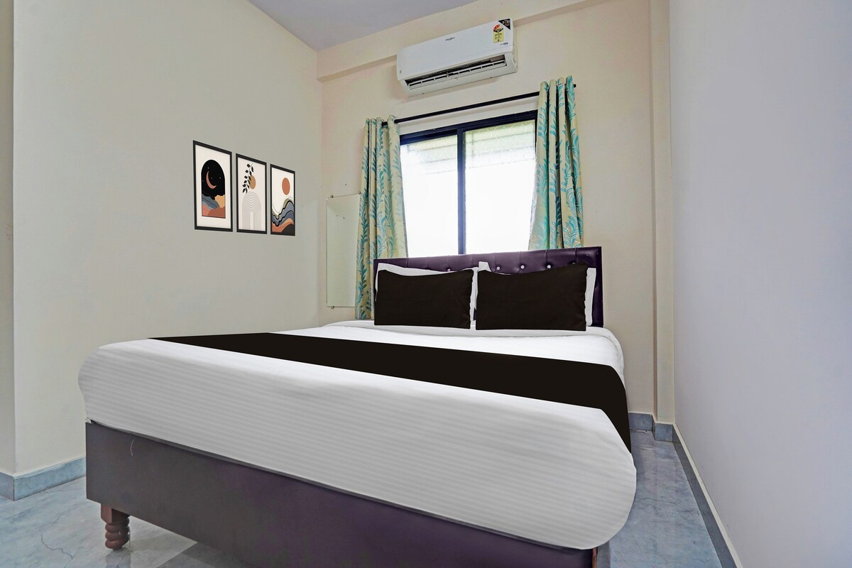 A hotel room with a bed, an air conditioner, a window with curtains, and framed artwork on the wall.