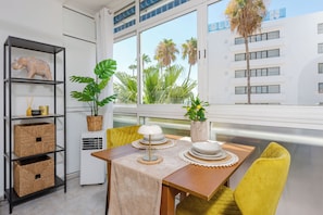 A dining area with a table set for two, a bookshelf with decorative items, and a view of palm trees through large windows. - Al Andalus Torremolinos – Bright Coastal Apartment near La Carihuela, Restaurants & Centre (Torremolinos)