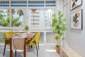 Een eetruimte met een tafel voor vier personen, gele stoelen, een potplant en uitzicht op palmbomen en een gebouw door grote ramen.