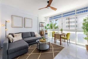 Una sala de estar moderna con un sofá gris, una mesa de centro y un ventilador de techo. Hay dos cuadros en la pared, una estantería con adornos, y vistas a un balcón con plantas.