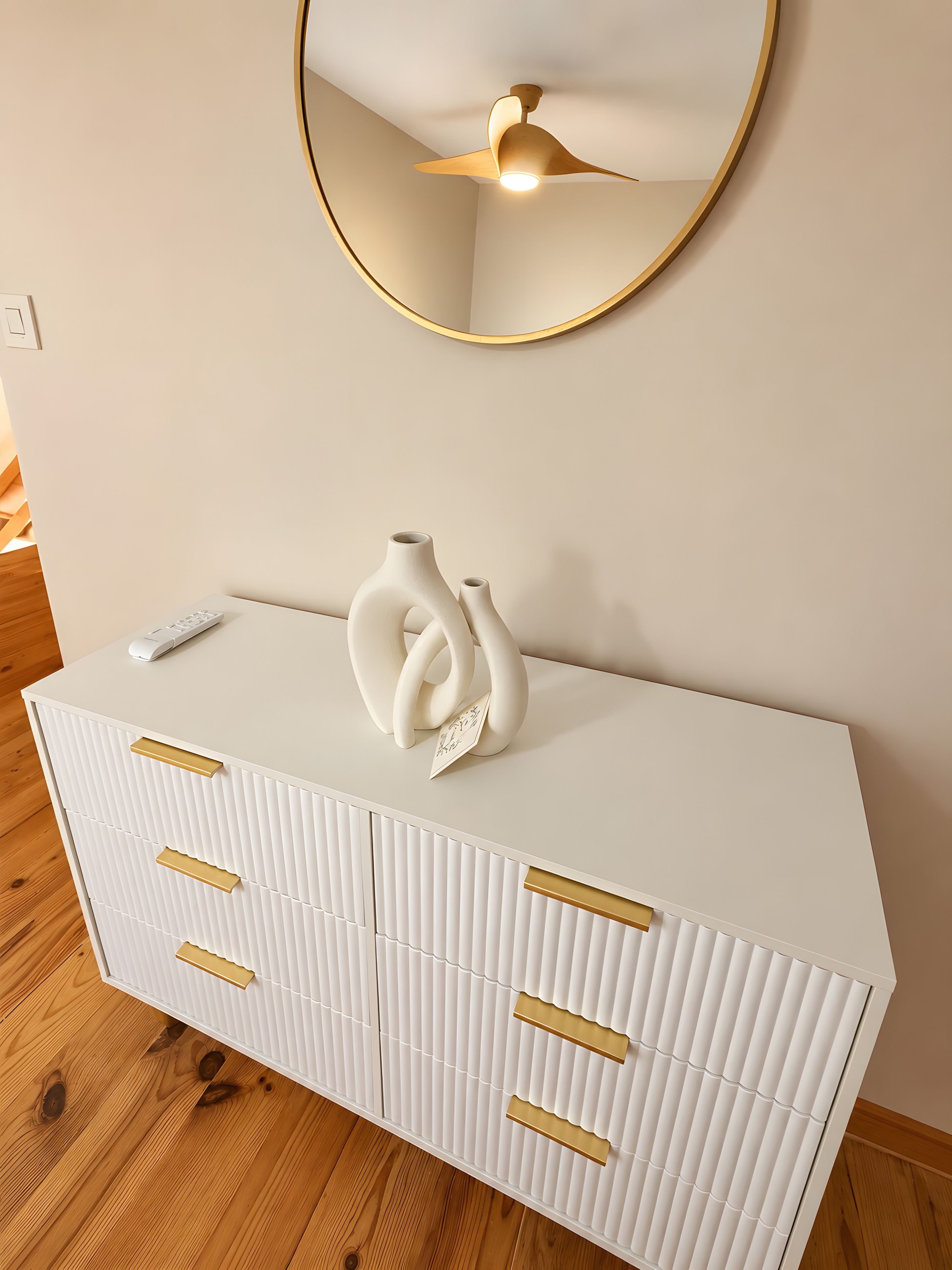 Ein weißes Schränkchen mit goldenen Griffen, eine weiße Vase und eine Fernbedienung darauf.