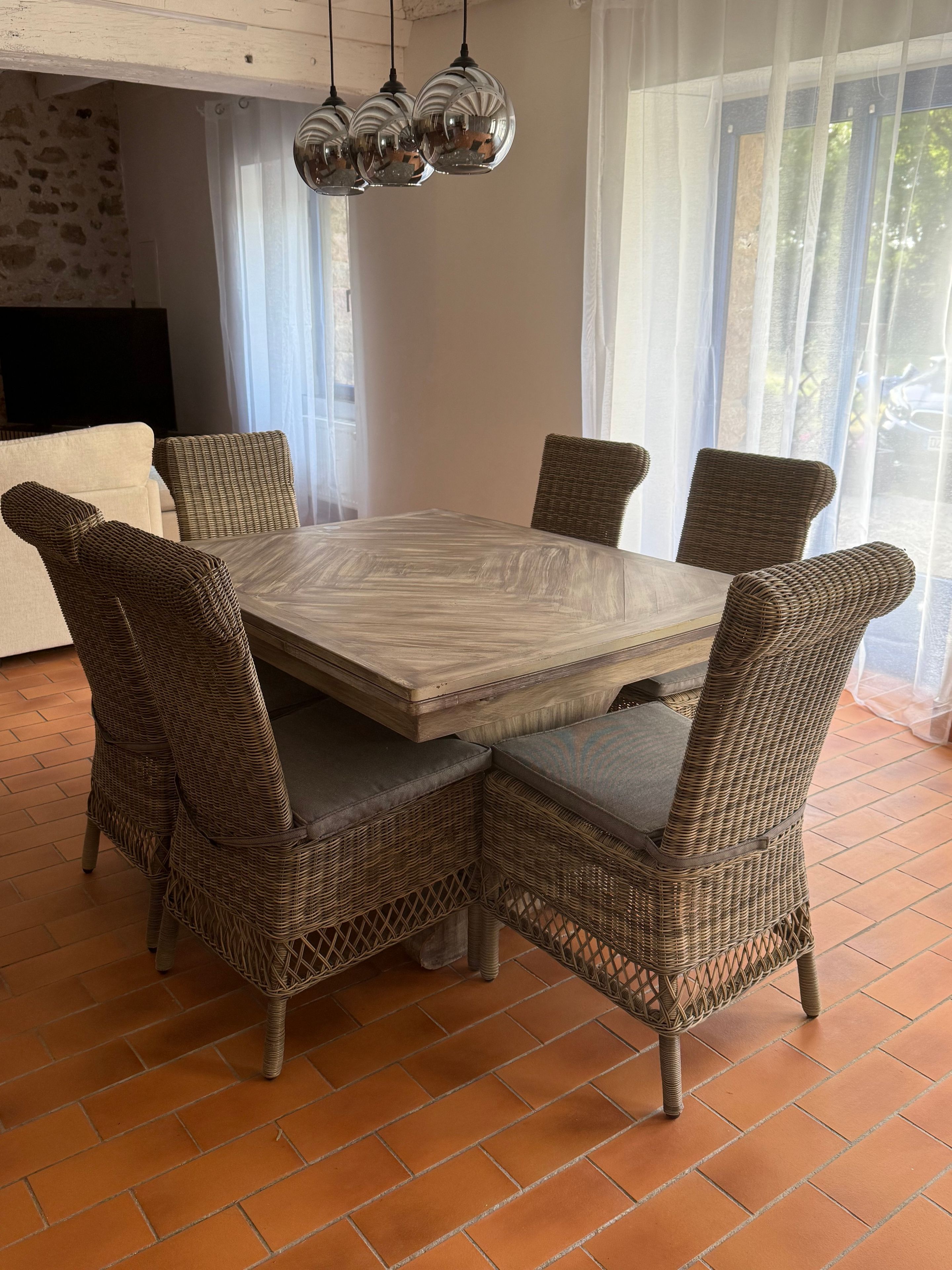 Un coin repas avec une table carrée entourée de chaises en osier, un luminaire suspendu au-dessus et une vue sur l’extérieur grâce à de grandes fenêtres.