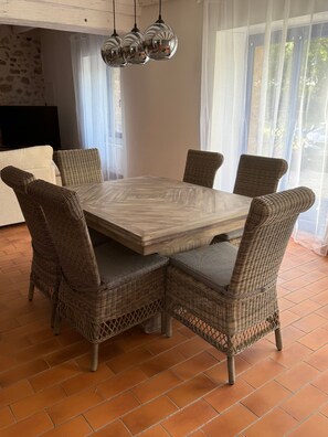 Un coin repas avec une table carrée entourée de chaises en osier, un luminaire suspendu au-dessus et une vue sur l’extérieur grâce à de grandes fenêtres.