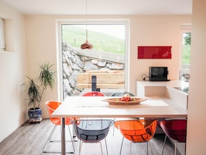 A modern dining area with a white table, orange chairs, and a view of a stone wall and greenery outside. - Chesa Montana - Top 101 (Maria Alm am Steinernen Meer)